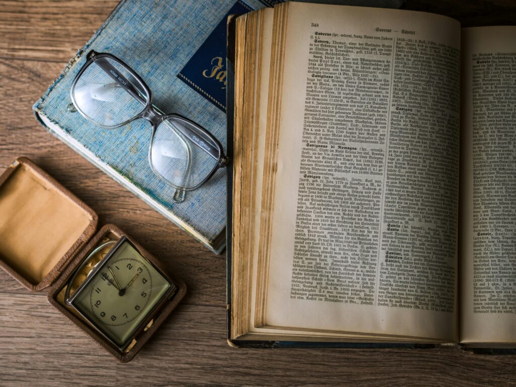 A vintage setting with antique books, watch, and reading glasses on a wooden table.